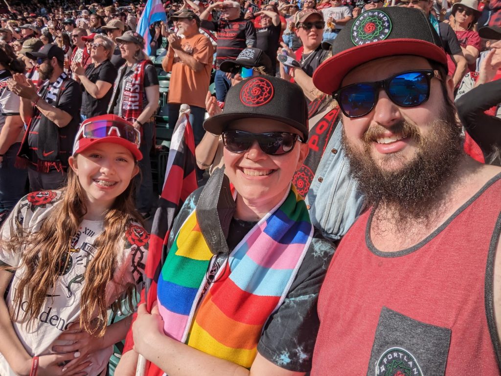 Photo of Sean, a bearded white man wearing a hat and sunglasses, with his wife and daughter at a Portland Thorns game