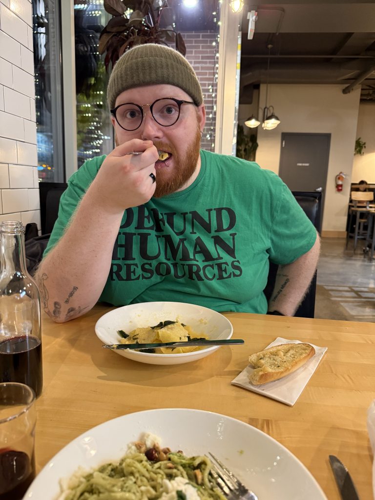 IMG_5142 Image of CG Brothers, a bearded white nonbinary person eating pasta wearing a t-shirt that says "defund human resources"
