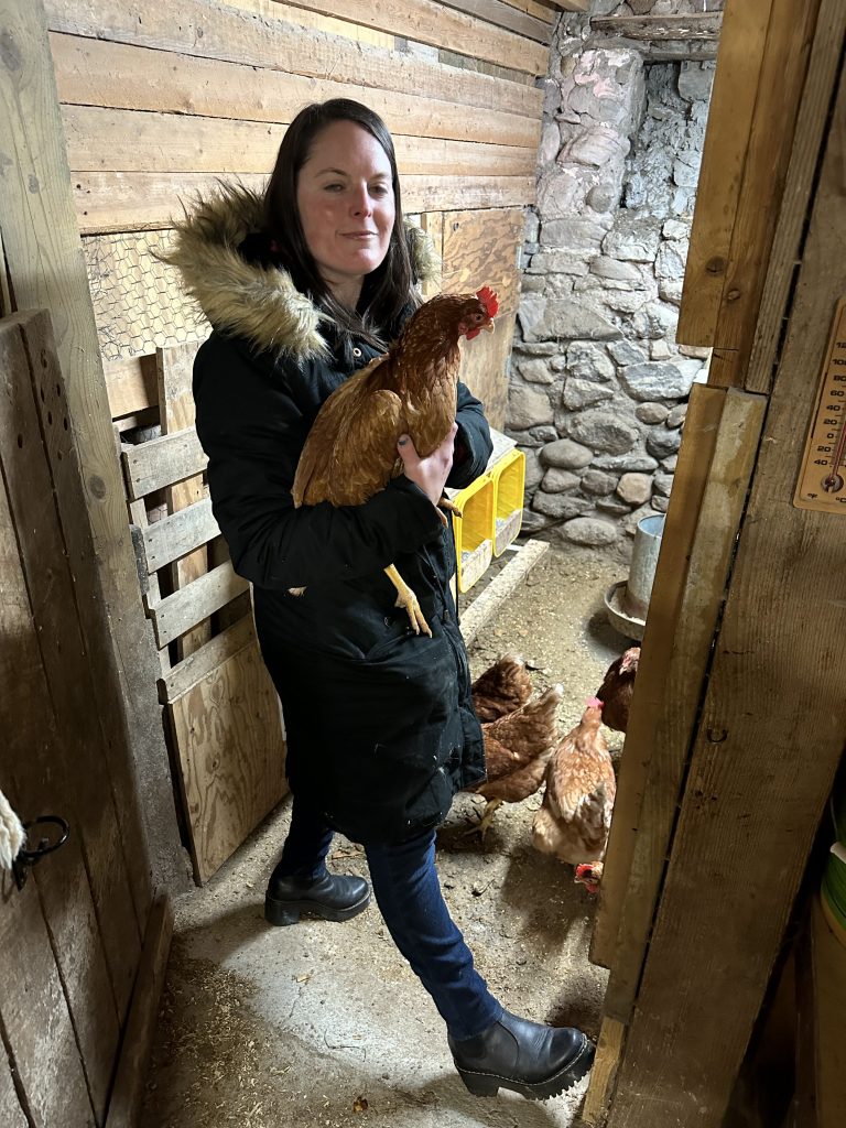 IMG_8874 Image of Brianna, a white woman with long brown hair wearing a three-quarter length winter coat and holding a chicken